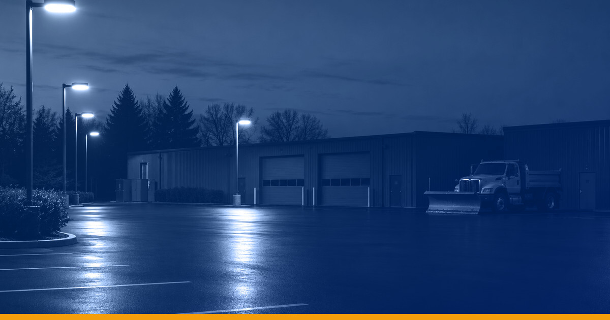 A freshly sealed parking lot at dusk with a parked snowplow and lit maintenance bays, pole lights at the upper left, under a Kardin navy gradient overlay.